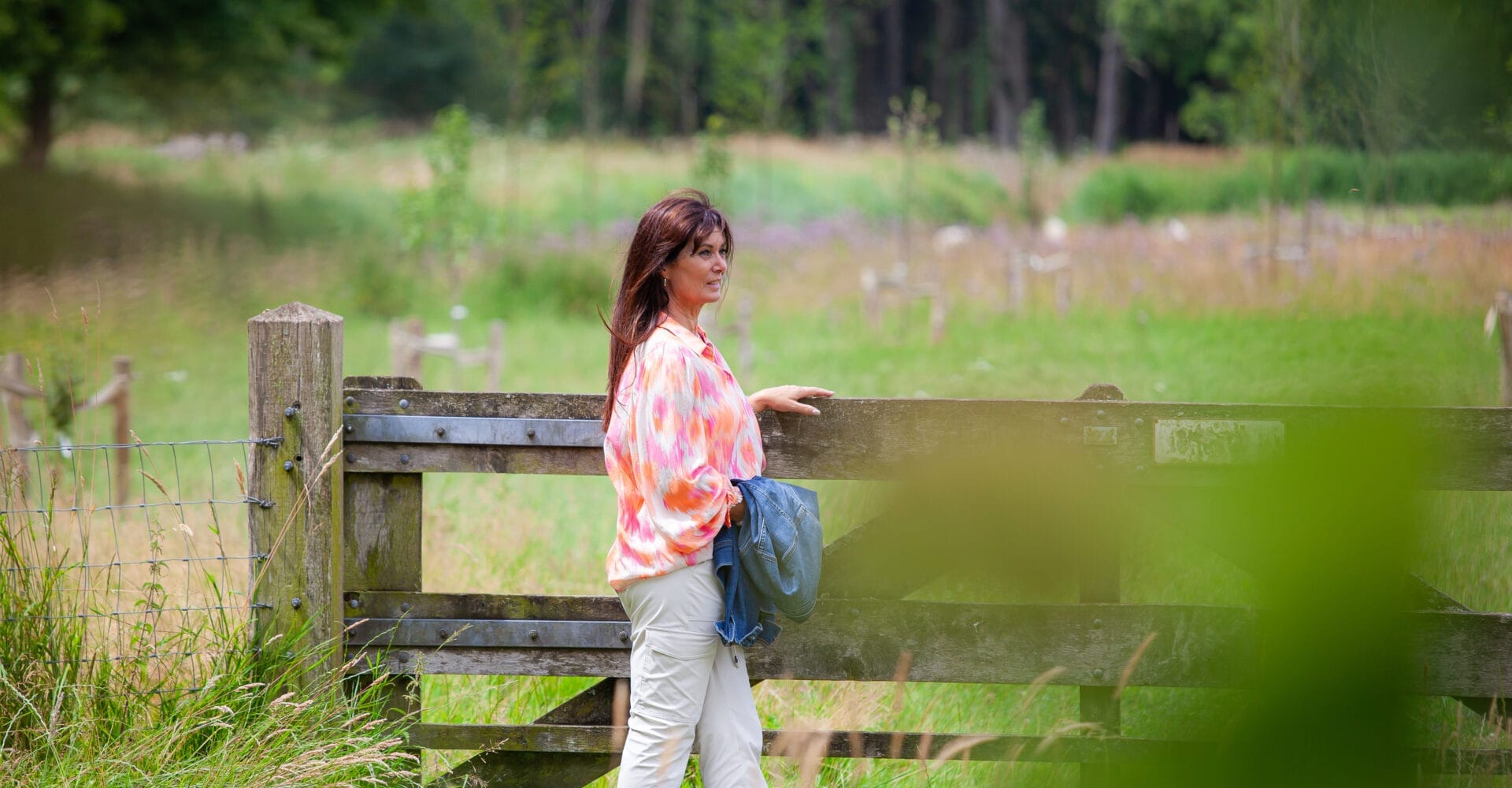 Een vrouw in een kleurrijke blouse en lichte broek staat bij een houten hek in een weelderig grasveld met bomen op de achtergrond. Ze houdt een jas vast en kijkt in de verte.